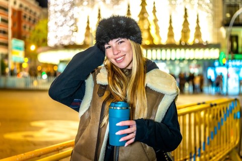Stylish woman holding reusable cup with hot drink, enjoying magical christmas lights and festive