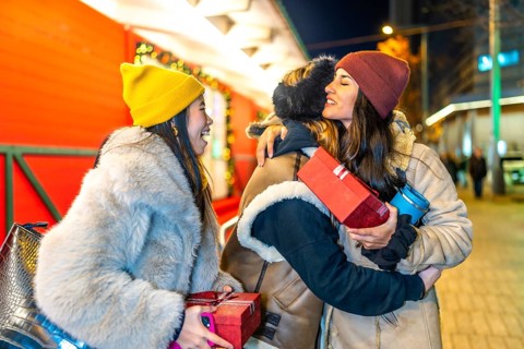 Three female friends exchanging christmas presents and hugging at a christmas market at night