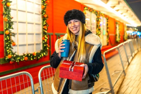 Young woman enjoying a warm beverage and holding a christmas present at a festive night market