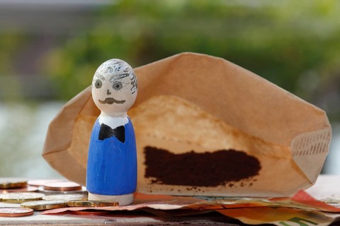 Male wooden cone figure with coins and banknote in front of filter bag with coffee North
