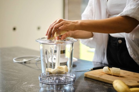 Sliced bananas are placed in a blender while a sunny kitchen provides a bright backdrop. Fresh