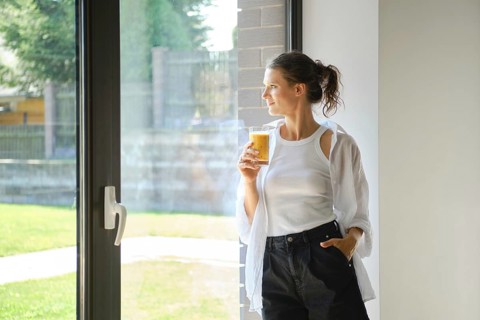 A woman in a white top and black pants stands by a window, holding a glass of smoothies. She gazes