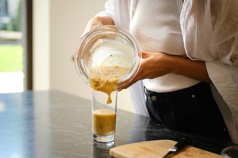Pouring a creamy banana and sea buckthorn smoothie from a glass bowl into a tall glass. The setting