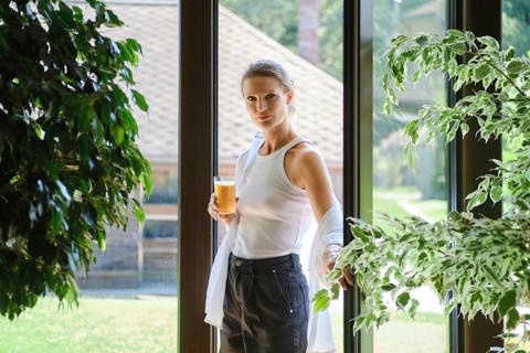 A woman stands by a large window, sipping a cool beverage while surrounded by green plants and
