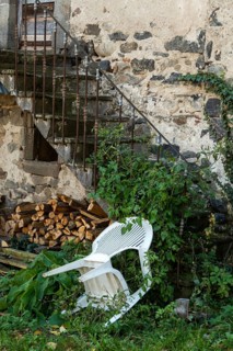 An unkempt outdoor area features a white chair leaning against a weathered stone wall, surrounded