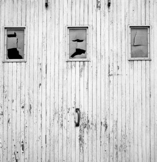 The weathered wooden wall, painted white, showcases three cracked windows that reveal a sense of
