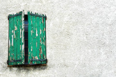 Weathered green shutters stand slightly ajar on a textured white wall, showcasing the charm of an