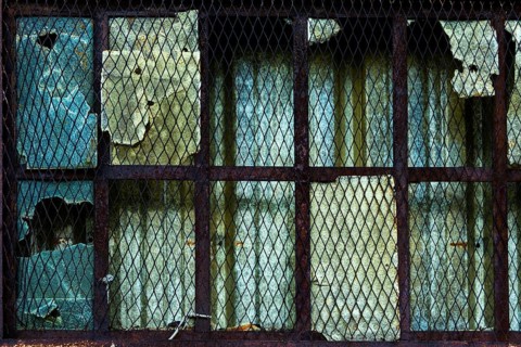 The weathered window features damaged glass and rusty metal bars, showing signs of decay and