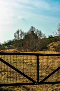 A picturesque view captures the ruins of a castle atop a hill, framed by a metal gate. The