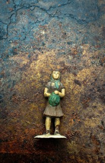 A small, weathered figurine depicting a girl in a dress, clutching a green bundle, stands on a