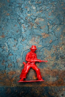 A small red plastic toy cowboy stands firmly on a rustic blue and brown textured background. The