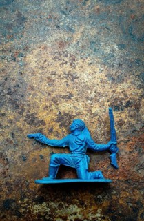 A blue plastic toy cowboy is kneeling, holding a pistol in one hand and a rifle in the other,
