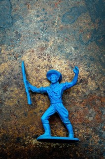 A small blue plastic toy cowboy stands firmly on a rustic blue and brown textured background. The