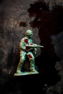 A green soldier figurine holds an assault weapon while displaying battle wounds marked by red
