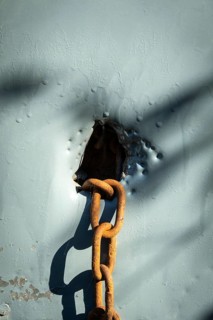 A rusty chain is situated through a weathered hole in a blue surface, with sunlight creating