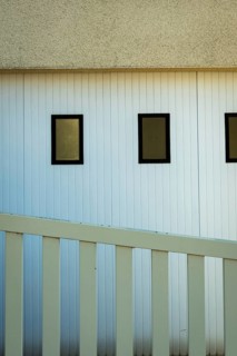 A building featuring three rectangular windows on a white vertical paneled wall. A white railing