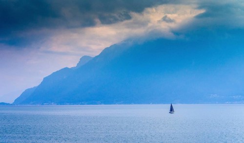 A lone sailboat navigates the tranquil waters of Lake Geneva surrounded by shadowy mountains. The
