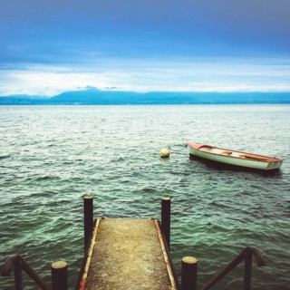 A wooden jetty reaches into the calm waters of Lake Geneva, where a small boat is moored. Mountains