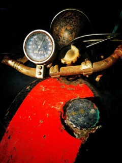 A worn motorcycle dashboard features a rusty speedometer and a faded paint job. The dark background