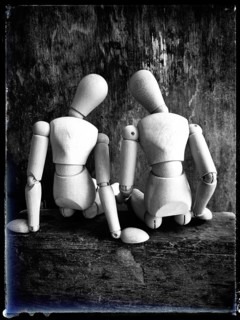 Two wooden figures are positioned closely together, facing away from the viewer. They sit on a