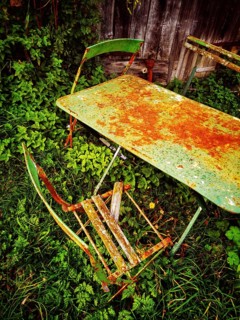 A rusty green table and a broken chair lay forgotten in thick, overgrown grass, evoking a sense of
