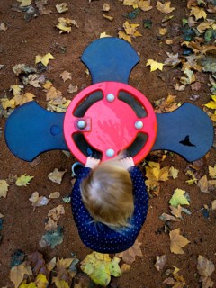 A young child is playing with a vibrant red spinner at a playground, surrounded by fallen yellow