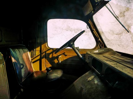 An abandoned truck interior features worn seats and an aged steering wheel, casting shadows against