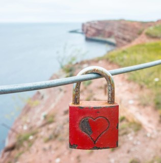 Red padlock with engraved heart symbol on a wire rope in front of the steep coast, red cliff of