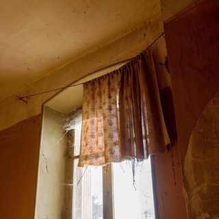 Sunlight streams through a dusty window adorned with worn curtains, highlighting the cobwebs in a