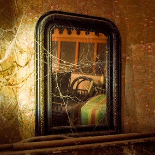 A weathered mirror stands in a neglected room, covered in cobwebs. The faded decor hints at past