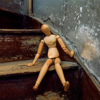 A wooden mannequin is seated casually on a weathered staircase, surrounded by rustic decor and