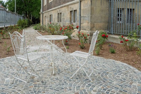 Two white metal chairs and a table on a paved square in front of a historic building, surrounded by