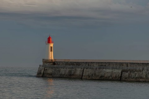 A lighthouse stands on a pier and casts its light over calm waters at dusk. The sky is filled with
