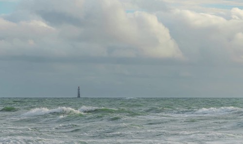Waves crash against the shore while a lone lighthouse looms in the distance under a sky filled with