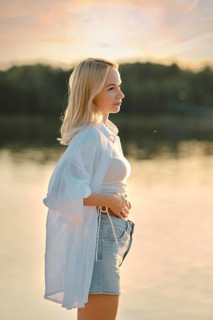 A young woman stands by a calm lake during sunset, feeling the gentle breeze. Her casual outfit