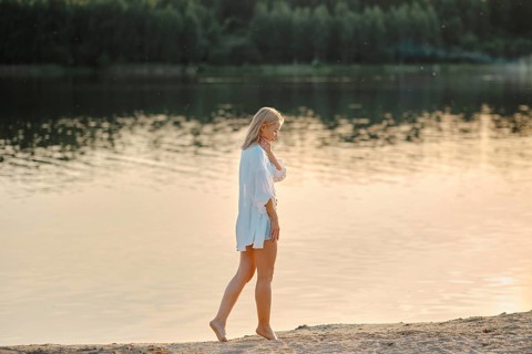 Pensive woman strolls barefoot on the sandy beach beside a serene lake. Golden sunlight reflects