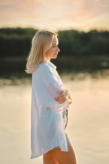 A young woman stands pensively by a calm lake at sunset, dressed in a light shirt and shorts. The