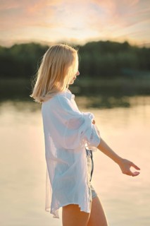 A young woman stands by the tranquil lake at sunset, wearing a white shirt and denim shorts. The