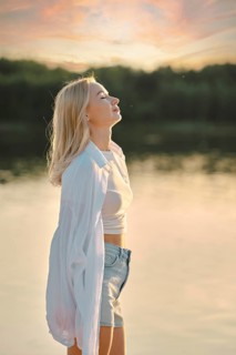 A woman stands by the calm lake, feeling the gentle breeze as she admires the colorful sunset sky.