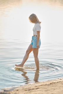 A woman stands in shallow water at a beach during golden hour, playfully splashing around with her
