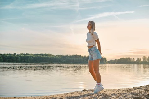 A young woman walks leisurely along the sandy lakeshore as the sun sets behind the trees. She wears