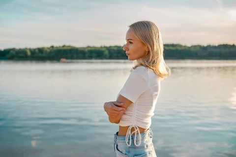 A blonde woman stands in profile by a calm lake, gazing thoughtfully into the distance. The sun