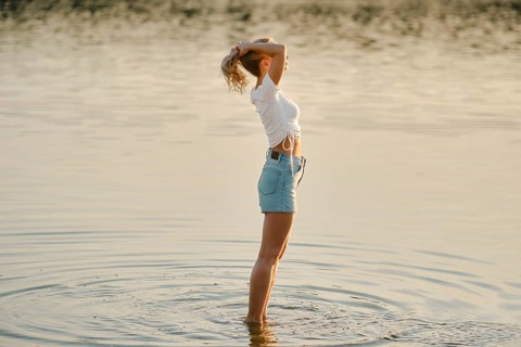 A woman stands in shallow water in profile, basking in the warm glow of sunset. She raises her