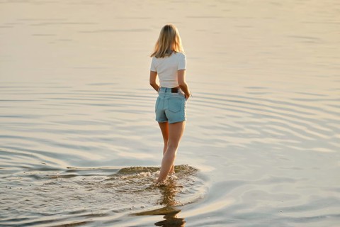 A woman with blonde hair wades through calm, shallow water as the sun sets behind her, creating a