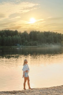 A woman strolls barefoot along the sandy shore of a serene lake during sunset. The sun sets behind