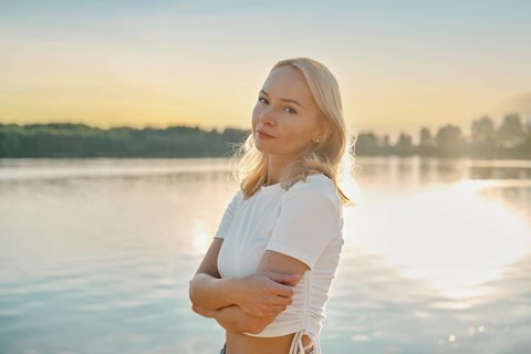 Cute woman stands thoughtfully by the lake, gazing into the distance as the sun sets behind her.