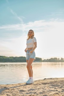 A young woman stands on a sandy beach, facing the camera. She wears a casual outfit of a cropped