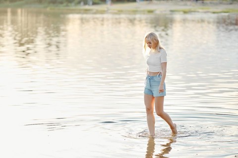 A young woman with blonde hair strolls through shallow water in a lake during sunset. The bright