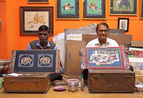 Miniature painters, aged 50 and 52, show paintings of elephant and royal city palace in studio,