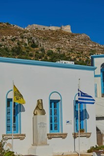 Bust in front of a white building with blue windows, Greek flag and yellow flag in the foreground,